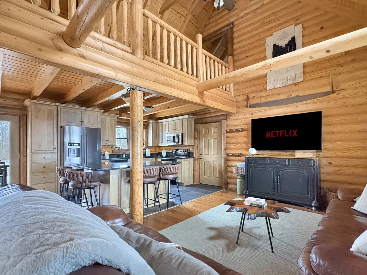 Interior of a cozy log cabin with a wood-paneled living room and kitchen. The living room has a leather sofa, a TV displaying the Netflix logo, and a small wood coffee table. The kitchen features light wood cabinets, stainless steel appliances, and a