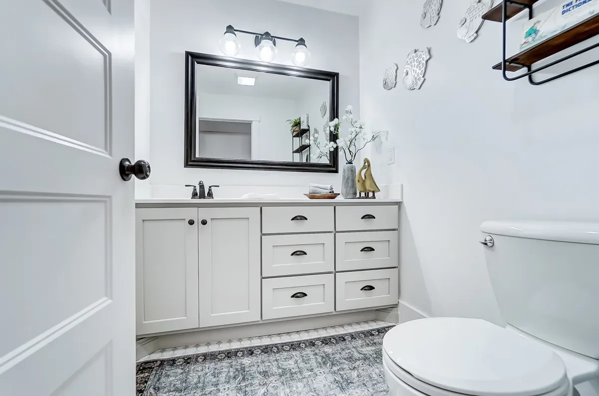 A modern white bathroom with a white vanity, black hardware, a large mirror, decorative wall arts, a vase with flowers, and a toilet.