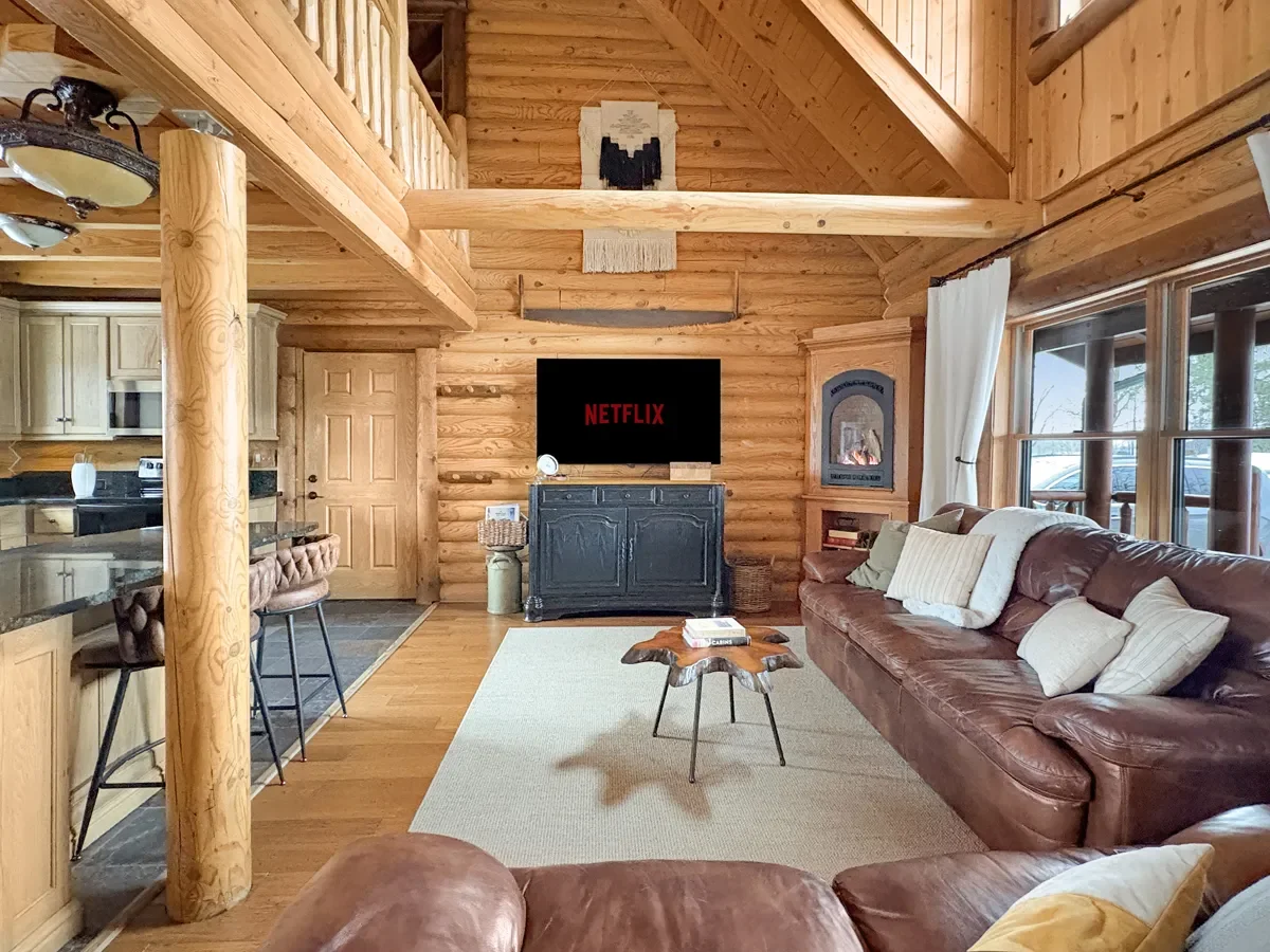Interior view of a cozy log cabin living room with wooden walls, a leather sofa with pillows, a flat-screen TV displaying Netflix, a wood-burning stove, a window with curtains, and a small coffee table on a light-colored rug.