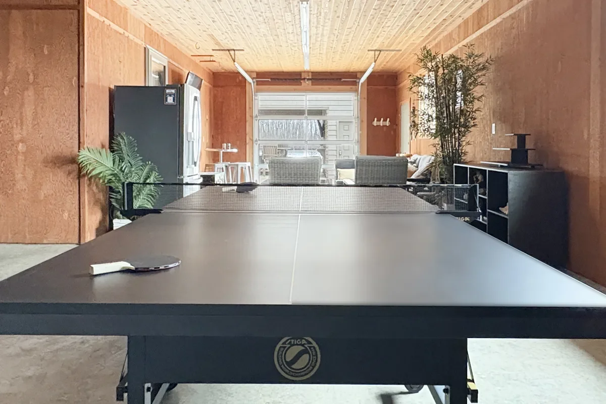 Indoor game room with a ping pong table in the center, a refrigerator on the left, and a seating area with chairs and a sofa in the background.