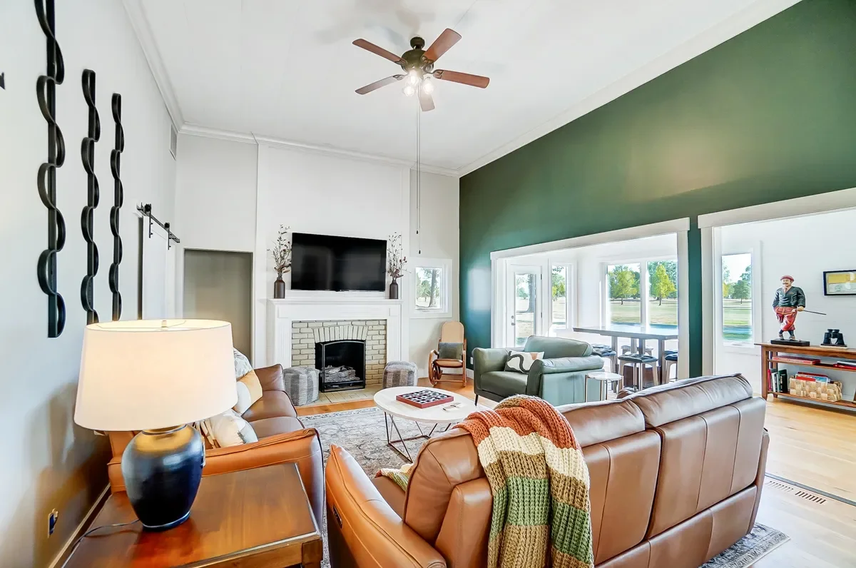 Living room with a green accent wall, a white brick fireplace, a flat-screen TV above fireplace, in front of a large window with a sitting area, brown and green furniture, a coffee table with a game, and a lamp on a side table, hardwood floors, ceili