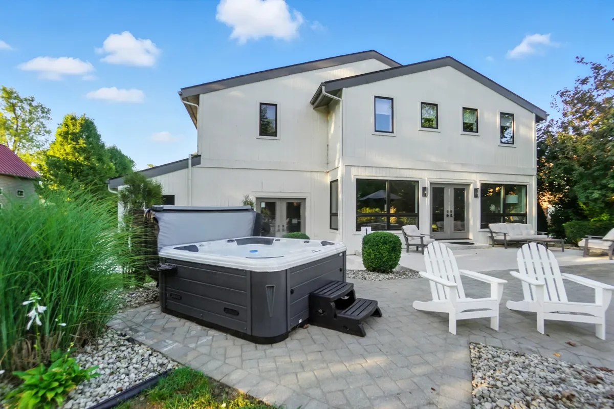 Backyard patio with mobile hot tub, white Adirondack chairs, and outdoor seating area with a two-story house in the background, surrounded by trees.