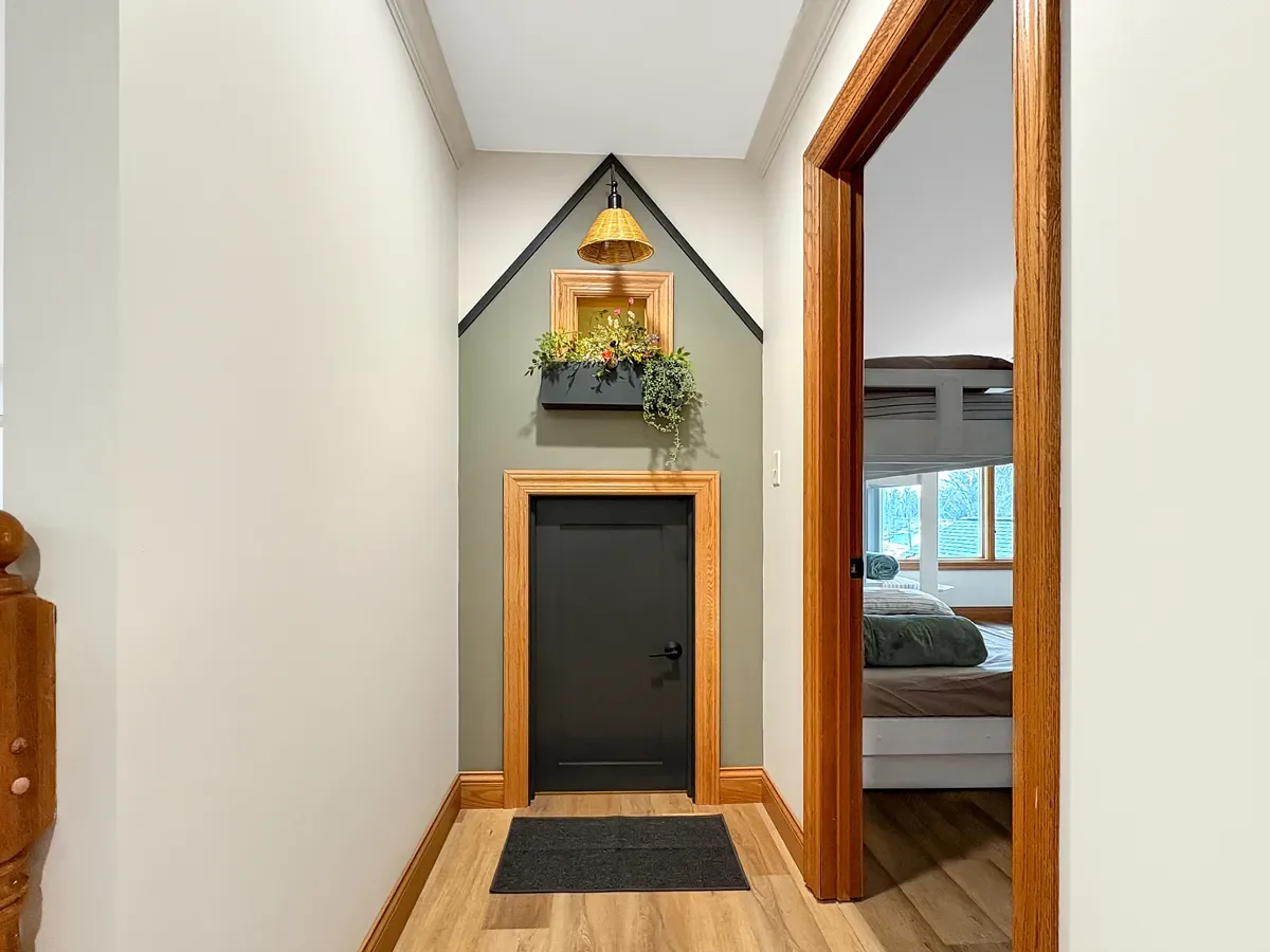 A hallway with a small black door, a vaulted ceiling decorated with a hanging light fixture, a wooden framed window with flower decoration, and a doorway leading to a bedroom with a bed and windows showing a snowy landscape outside.