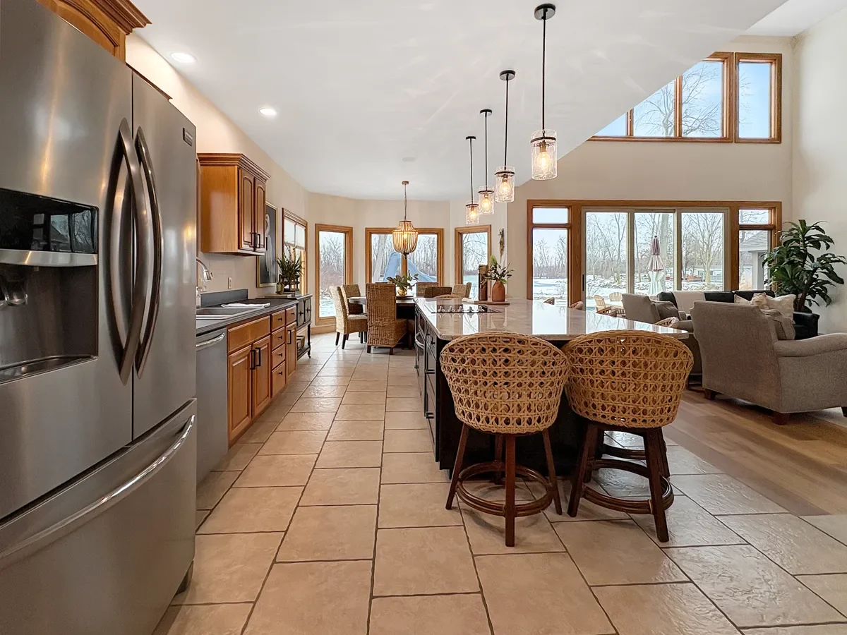Open-concept kitchen and living room area with large windows showing an outdoor view, featuring a kitchen with wooden cabinets, stainless steel refrigerator, tiled floor, island with bar stools, and a living area with sofas and chairs.