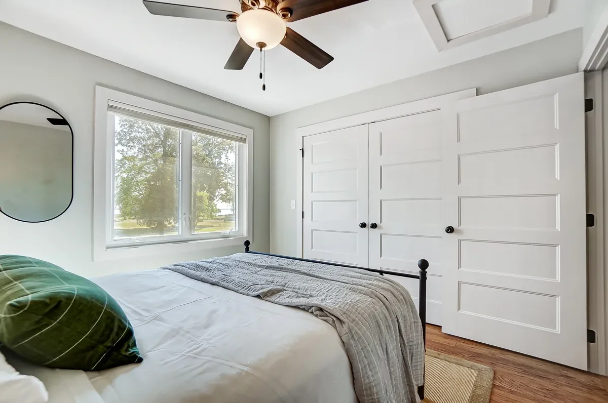 A bedroom with a window showing trees outside, a white bed with a green pillow and a gray blanket, a closet with sliding white doors, a ceiling fan with dark blades, a mirror on the wall, and wooden flooring.
