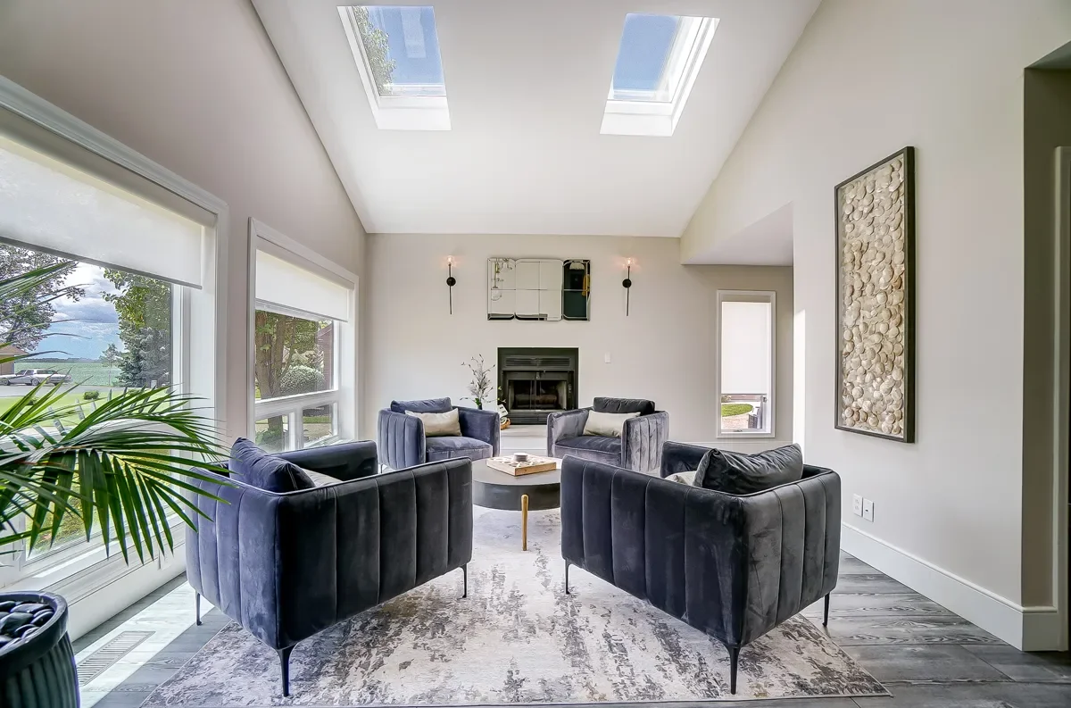 Living room with four navy velvet armchairs arranged around a coffee table, a fireplace with a mirror above it, large windows, and skylights providing natural light.
