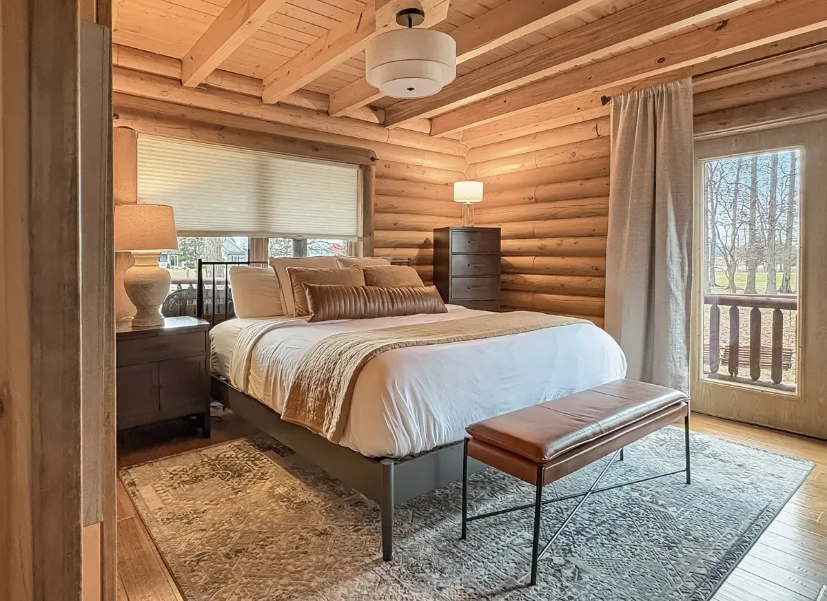 A cozy bedroom with log cabin walls, a bed with white bedding and brown pillows, a bench at the foot of the bed, nightstands with lamps, a chest of drawers with a lamp, a window with blinds, and a glass door leading to a balcony with a view of trees.
