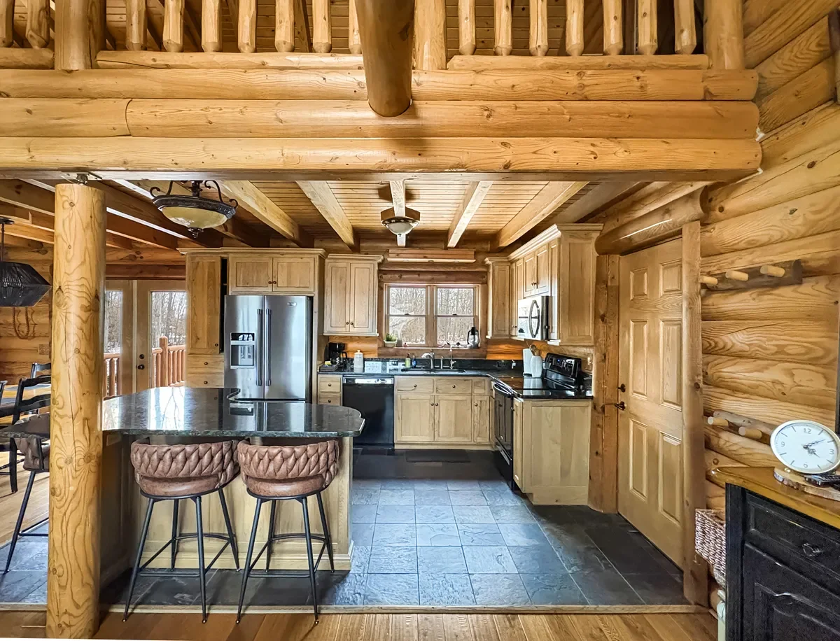 Kitchen in a log cabin with wooden walls, ceiling, and cabinets. Features stainless steel refrigerator, black appliances, and a window above the sink. There is a small counter with two padded chairs, and the floor is made of large gray tiles.
