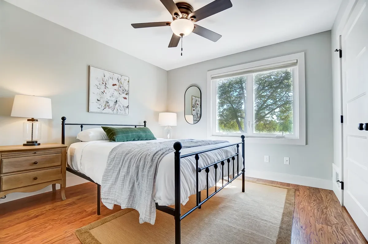 A bedroom with a black metal bed frame, white bedding, green pillows, and a gray blanket. A wooden nightstand with a lamp on each side of the bed. A large window showing trees outside, a mirror on the wall, and a ceiling fan with a light.
