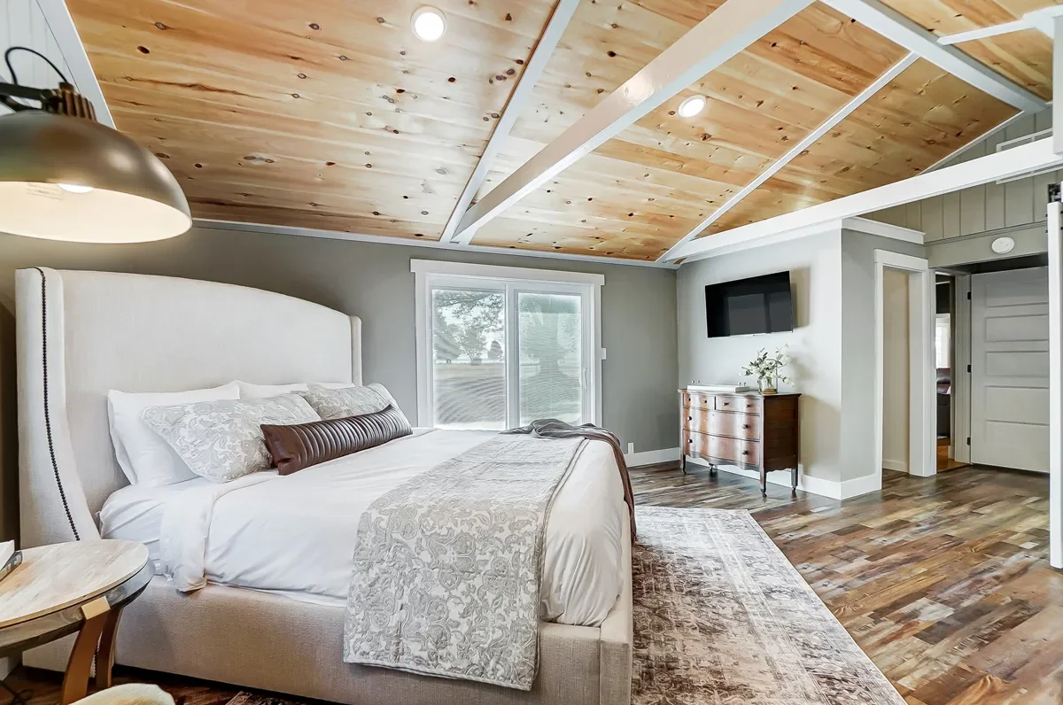 A bedroom with a large white bed, wooden ceiling, window, and hardwood floor.