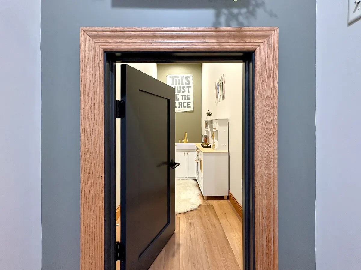 View through a doorway into a small room with a kitchenette, white cabinets, a gold faucet, and framed artwork on the walls.