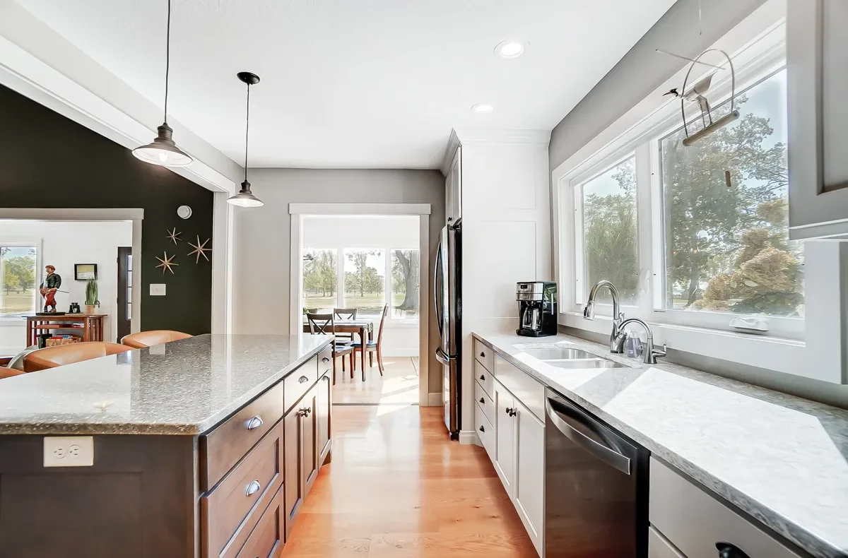 Bright kitchen with white cabinets, granite countertops, large window, and hardwood floors. A coffee maker and dishwasher are visible, with a view of a dining area and backyard outside.