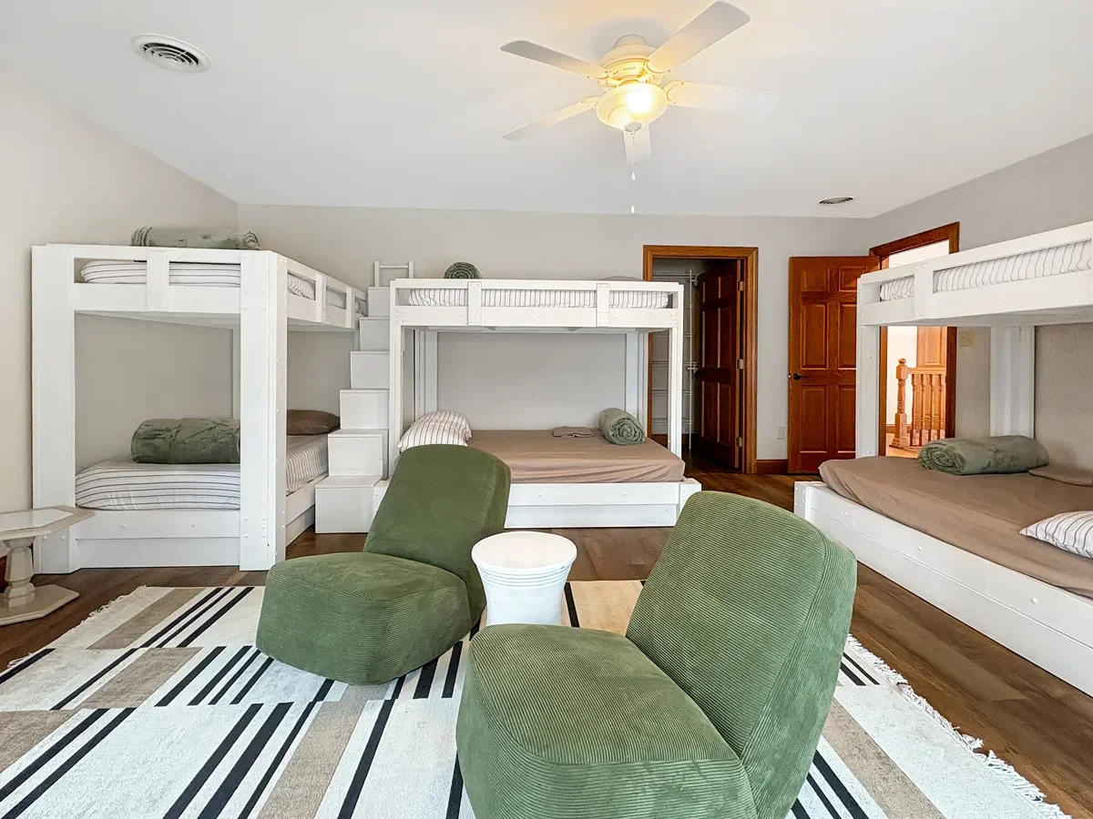 A bedroom with three sets of white bunk beds with mattresses and pillows, a ceiling fan with light, a door, a staircase with wooden railing, a patterned area rug, and two green chairs surrounding a small white table.