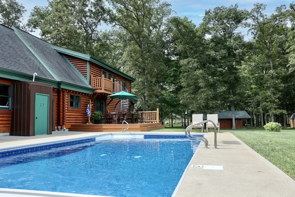 A backyard with a swimming pool, a wooden deck with outdoor furniture, and a log cabin-style house surrounded by trees.