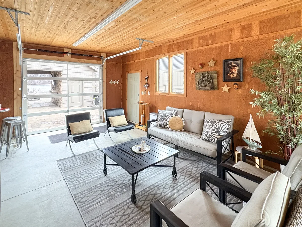 Living room with wooden walls and ceiling, featuring a glass garage door, a beige sofa with patterned pillows, two black chairs, a coffee table, a large potted plant, and wall decorations including framed pictures and star-shaped ornaments.