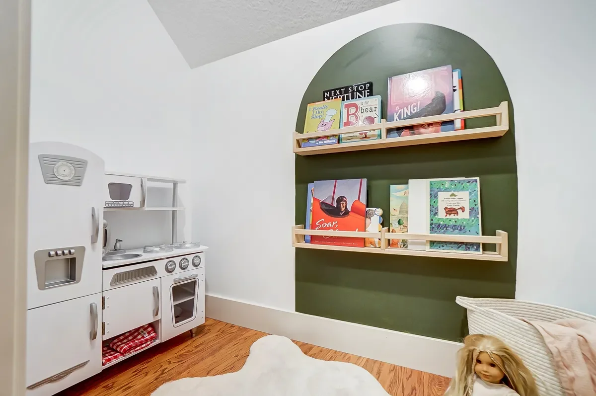 A children's playroom with a white toy kitchen set on the left, a green wall with two wooden shelves holding children's books on the right, and a doll in a wicker crib with a blanket on the bottom right.