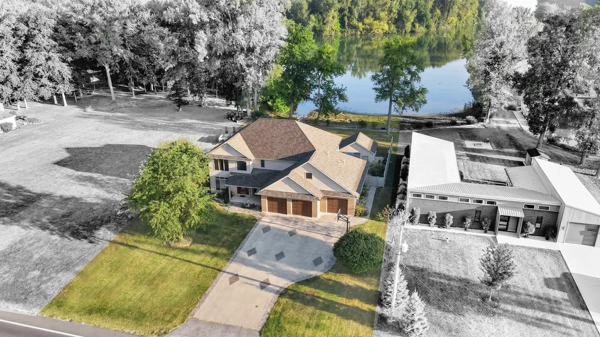 Aerial view of a neighborhood showcasing a house in color next to black and white houses, with a lake in the background and trees surrounding the area.