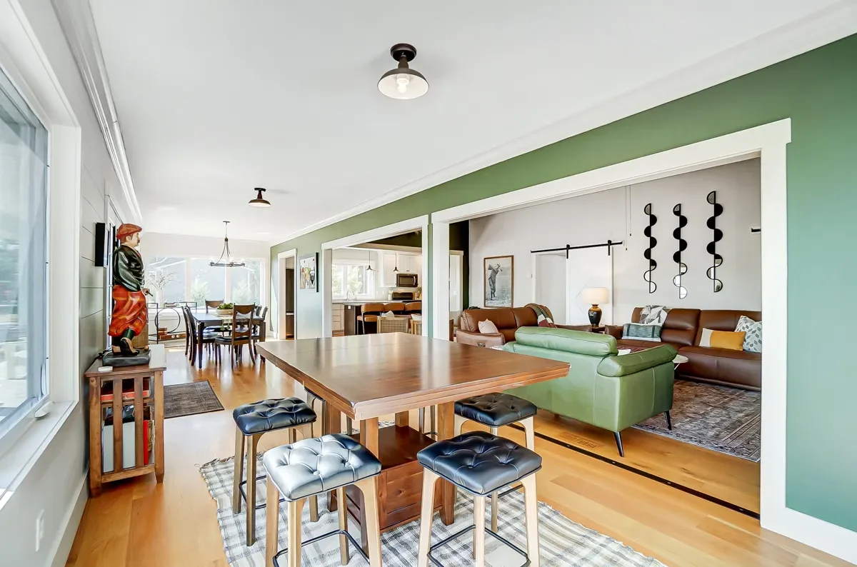 Open-concept living and dining room with green accent wall, wooden dining table and stools, brown couches, and a view of the kitchen.