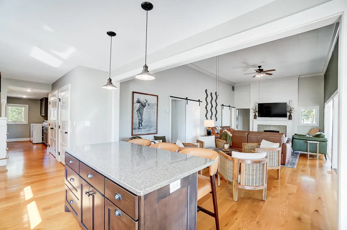 Open-concept living room and kitchen with hardwood floors, modern lighting, a brown leather sofa, armchairs, wall-mounted TV above a fireplace, and a kitchen island with barstools.