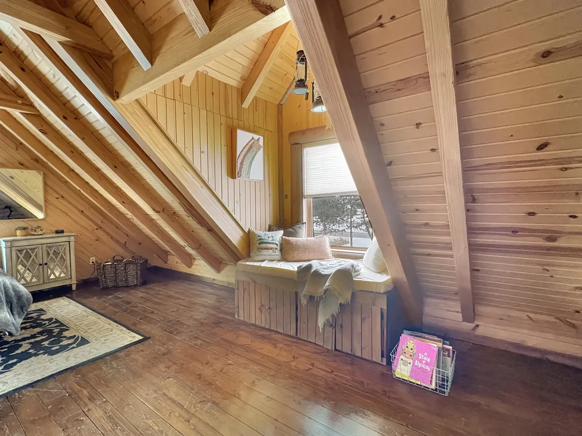 Wood-paneled attic room with sloped ceiling, window seat with pillows, and a bookshelf with magazines.