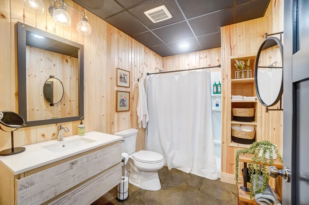 Bathroom with wooden wall paneling, a white sink with a mirror, a toilet, a shower with a curtain, and shelves with towels and plants.