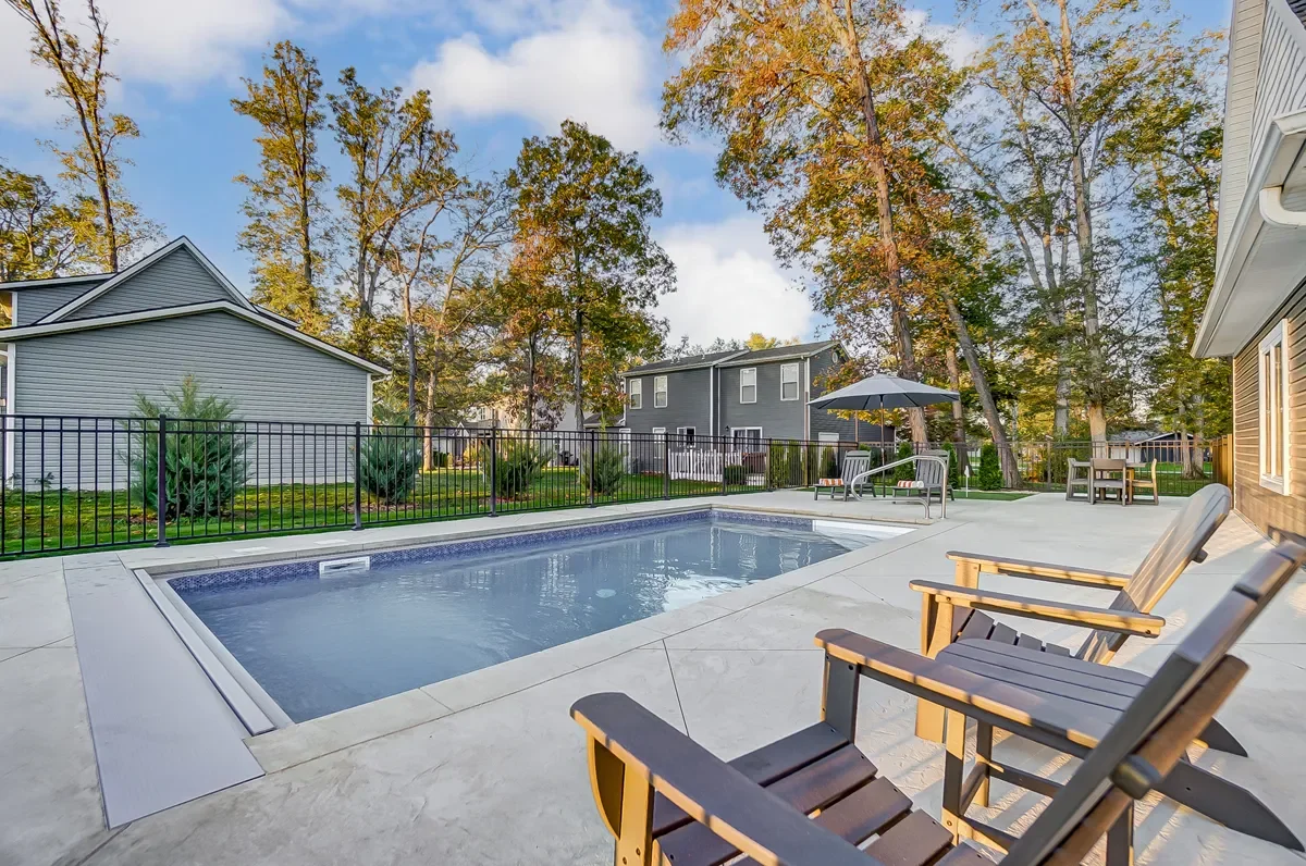Residential backyard featuring a rectangular in-ground pool, patio seating with wooden chairs, a patio table with an umbrella, surrounded by a black metal fence, with trees and houses in the background.