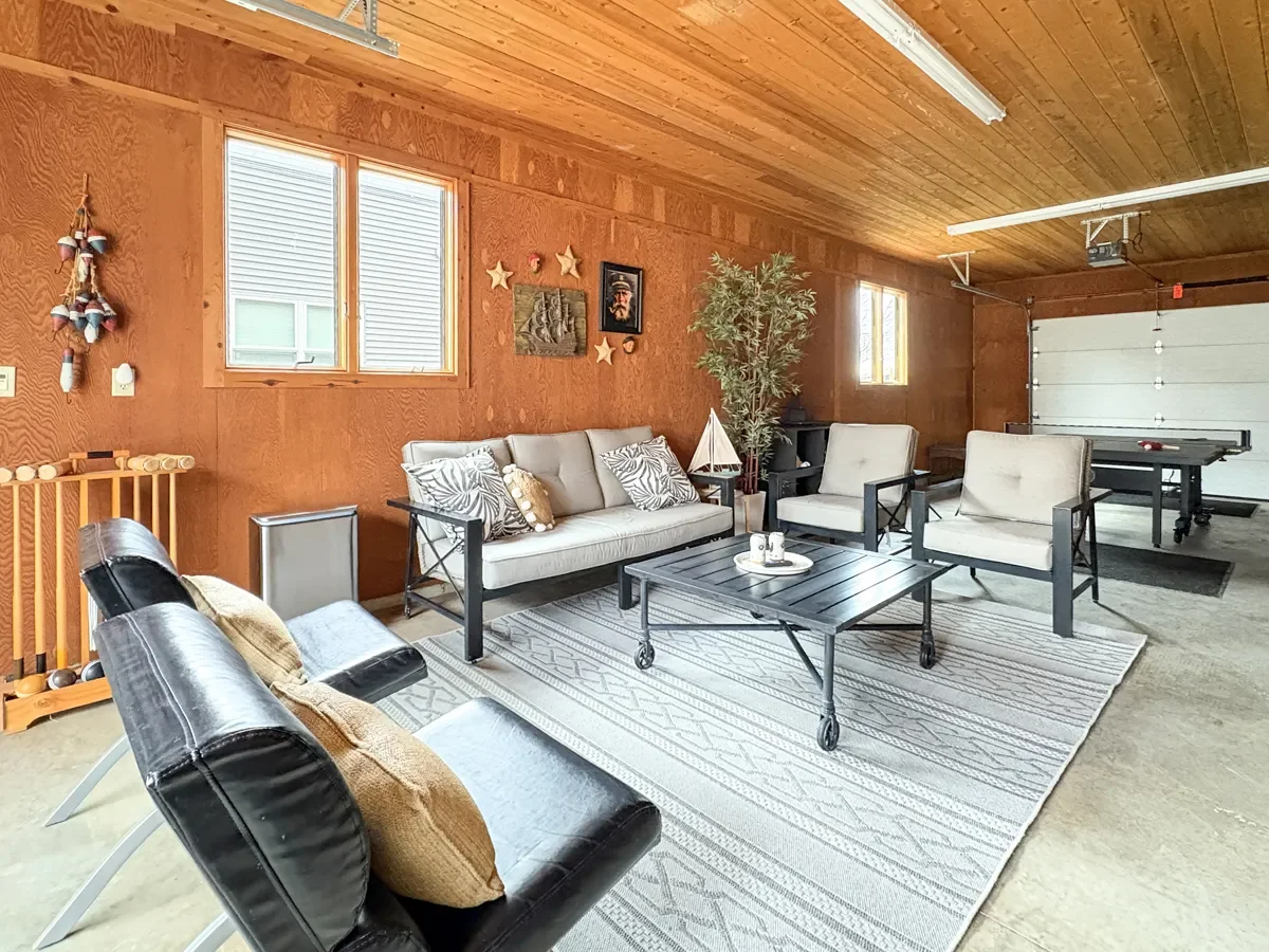 Living room with wooden walls and ceiling, furnished with a sofa, two armchairs, and a black leather chair. There's a coffee table on a patterned rug, decorative pillows, wall art, a tall plant, and windows letting in natural light. A foosball table 