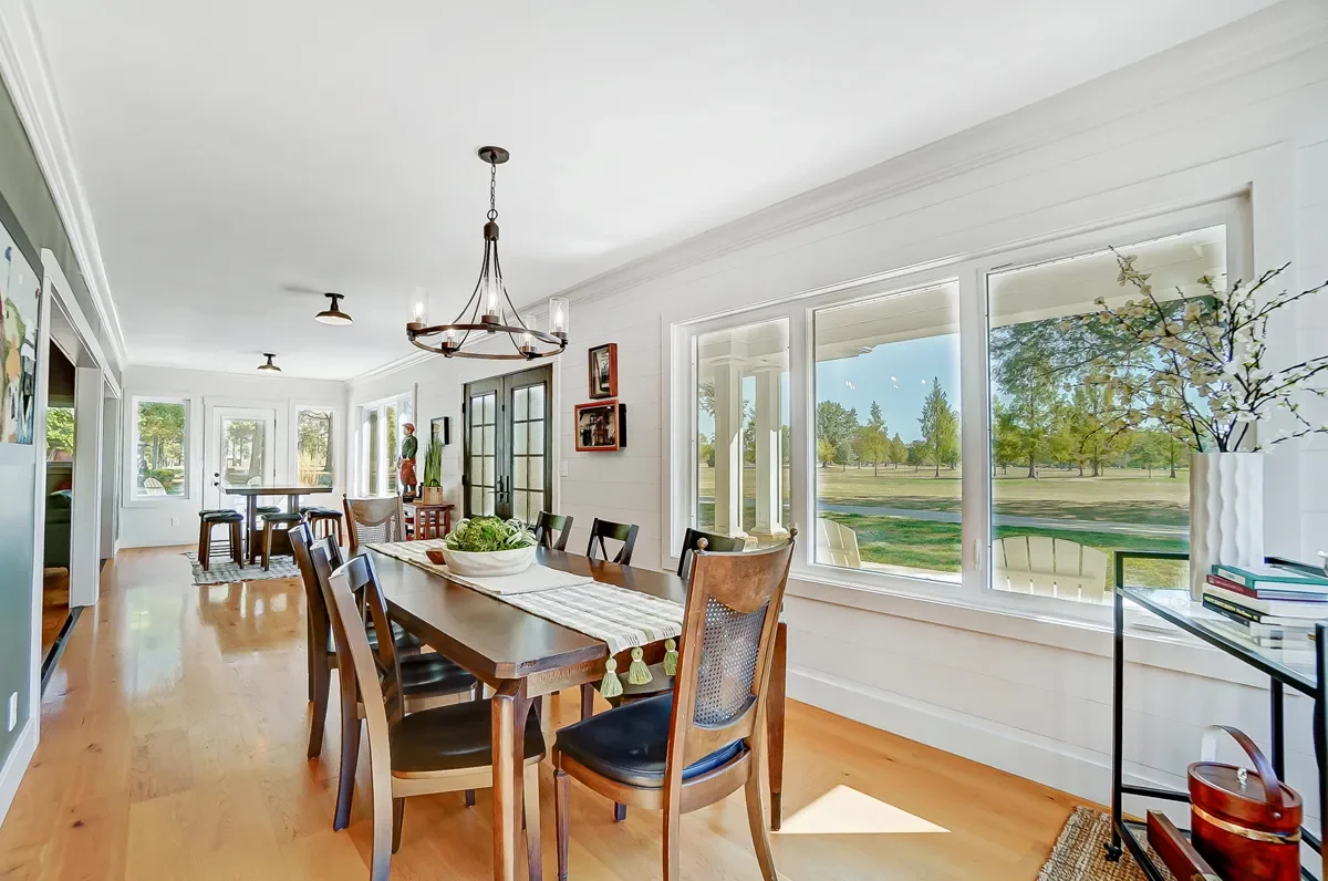 Bright dining room with large windows overlooking a grassy backyard, wooden dining table with six chairs, decorative chandelier, porcelain vase with branches, small black metal table with books, and an additional sitting area at the far end.