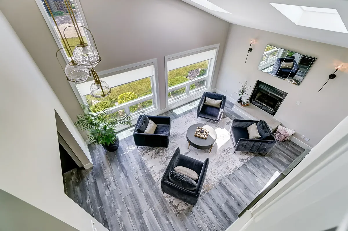 A modern living room with four black armchairs surrounding a round coffee table on a patterned rug, large windows letting in natural light, a fireplace with a mirror above it, a plant in a black pot, and a ceiling light fixture.