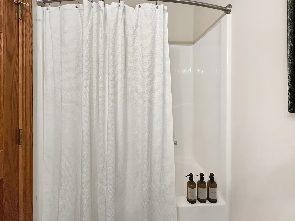 A white shower curtain in front of a shower stall with three brown bottles on a ledge inside.