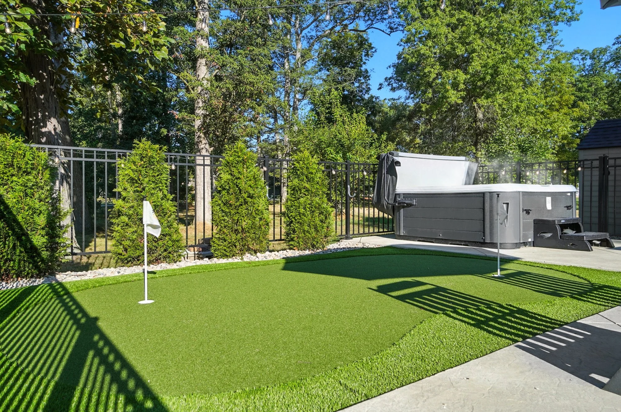 Backyard with artificial putting green, two small flags, and a hot tub against a black metal fence, surrounded by green bushes and trees under a clear blue sky.