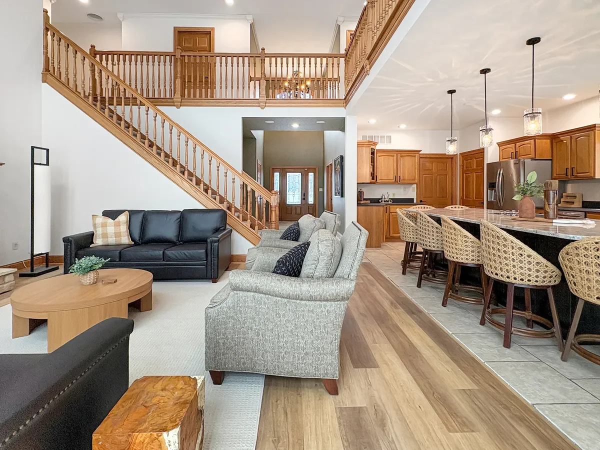 Open concept living room and kitchen with wooden staircase, black leather sofa, beige armchairs, wooden coffee table, rustic wood furniture, pendant lighting, and a kitchen with wooden cabinets, a large island with woven chairs, and stainless steel a