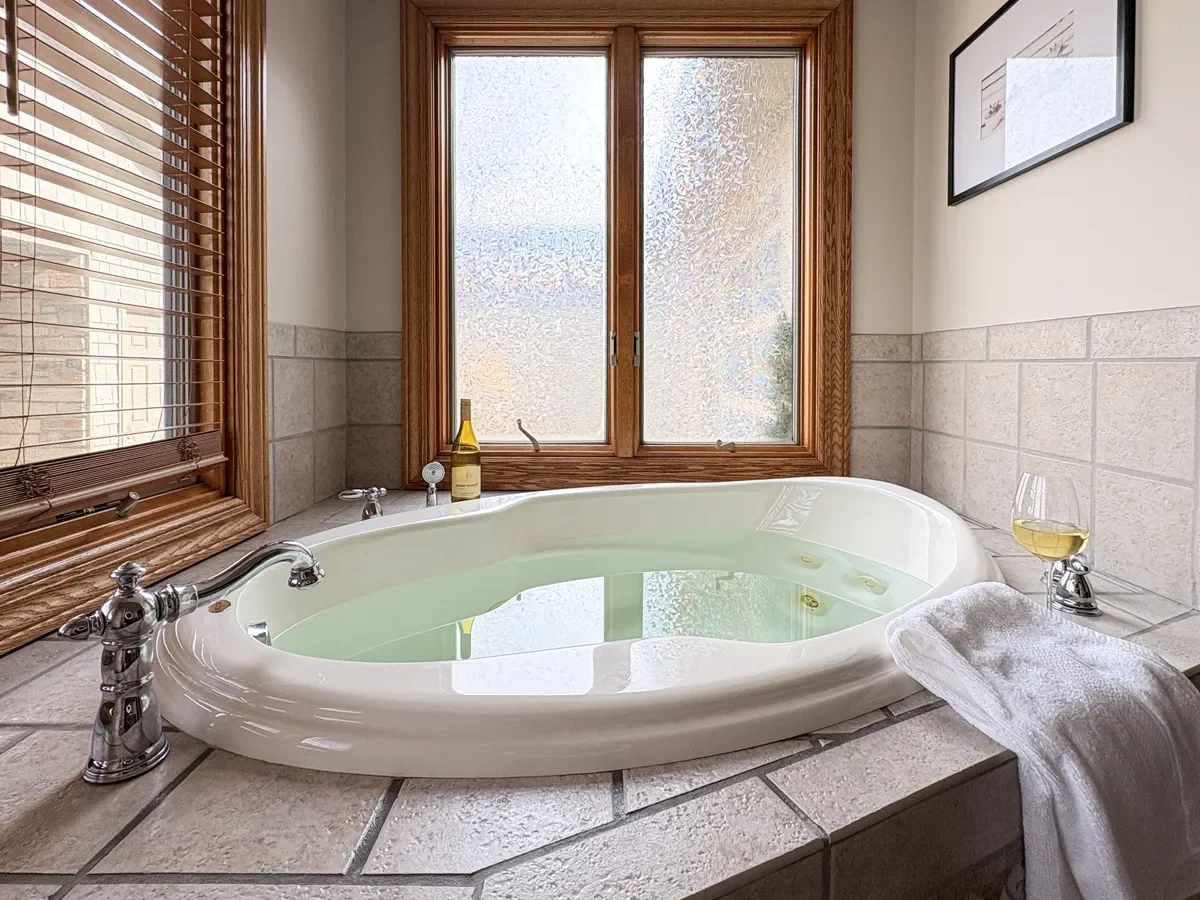 A bathroom with a corner jacuzzi tub filled with water, next to a window with frosted glass, surrounded by wooden trim and beige tile. On the tub's edge, there is a glass of white wine, a folded white towel, a bottle of wine, and a small white soap d