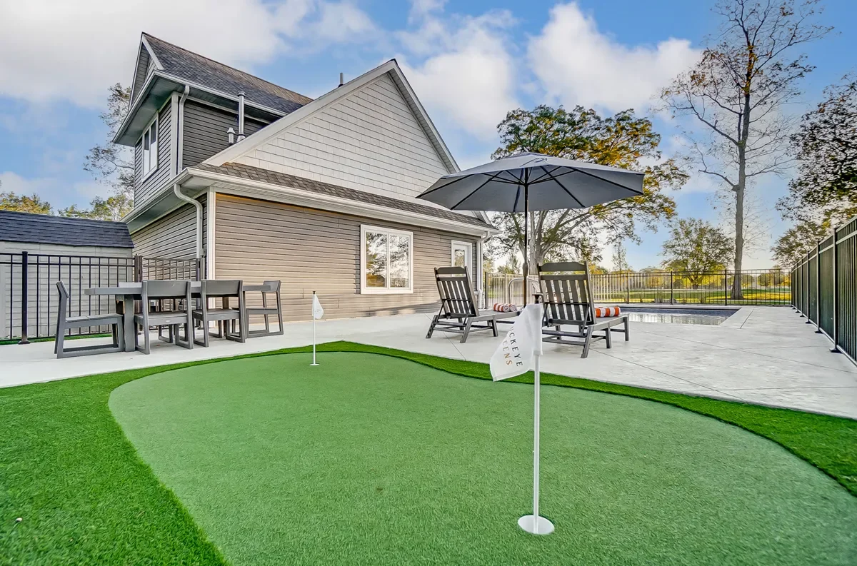 Backyard with putting green, outdoor furniture, umbrella, and house with grey siding in the background.