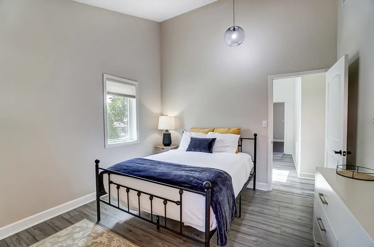 Bedroom with a black metal bed frame, white bedding, navy blue blanket and pillows, yellow headboard pillows, a white dresser, a nightstand with a lamp, a window with a shade, a ceiling light fixture, and a door leading to a hallway.