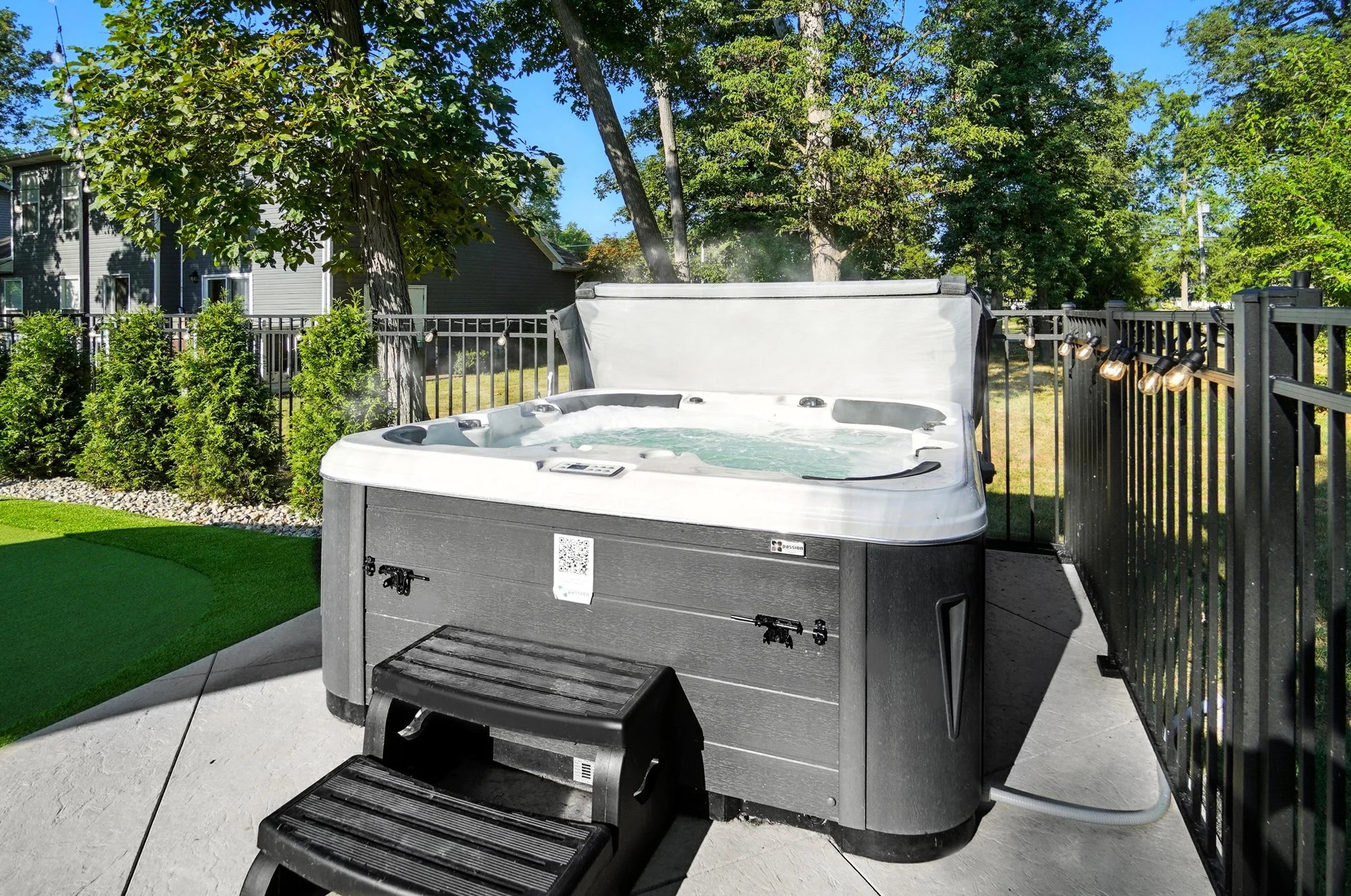 An outdoor hot tub with a white interior and dark gray exterior, placed on a concrete patio and surrounded by a black metal fence. The hot tub is steaming, indicating it is warm, and there is a small black step stool in front of it.