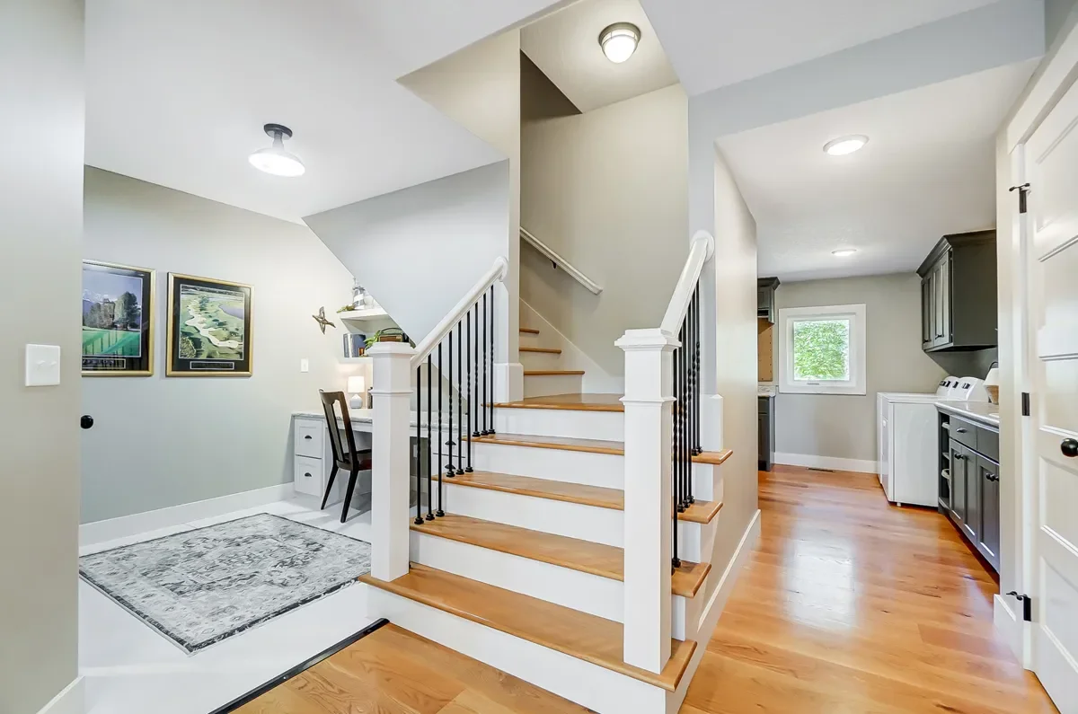 Interior of a house showing a staircase, a small desk with a chair, and a laundry area with washing machines and cabinets, with hardwood floors and light-colored walls.