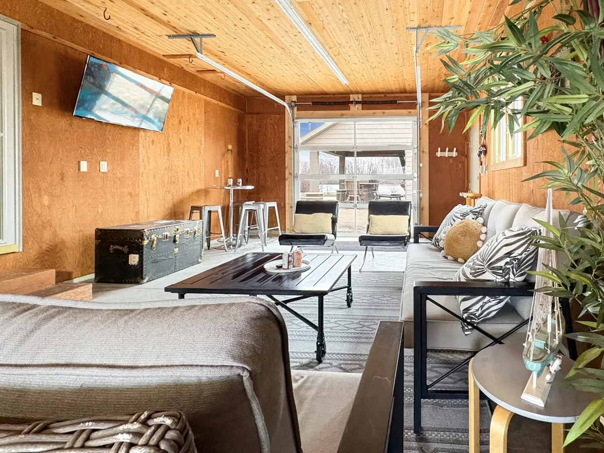 Living room with wood-paneled walls and ceiling, large front door with window, mounted TV, black trunk, bar table with stools, black chairs with beige pillows, striped rug, beige sofa with decorative pillows, side table with plants and decorative ite