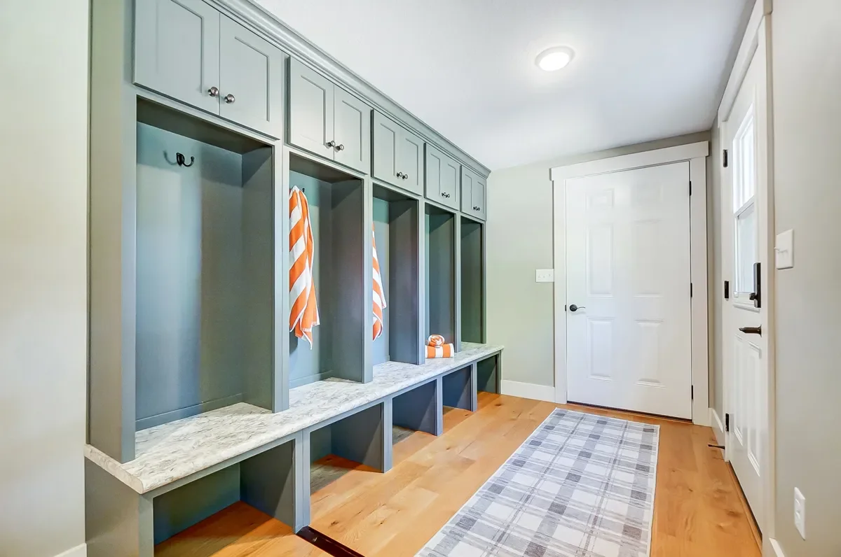 Mudroom with green built-in lockers, some with orange and white striped towels, a white marble bench, a gray plaid rug, and a white door in a house interior.