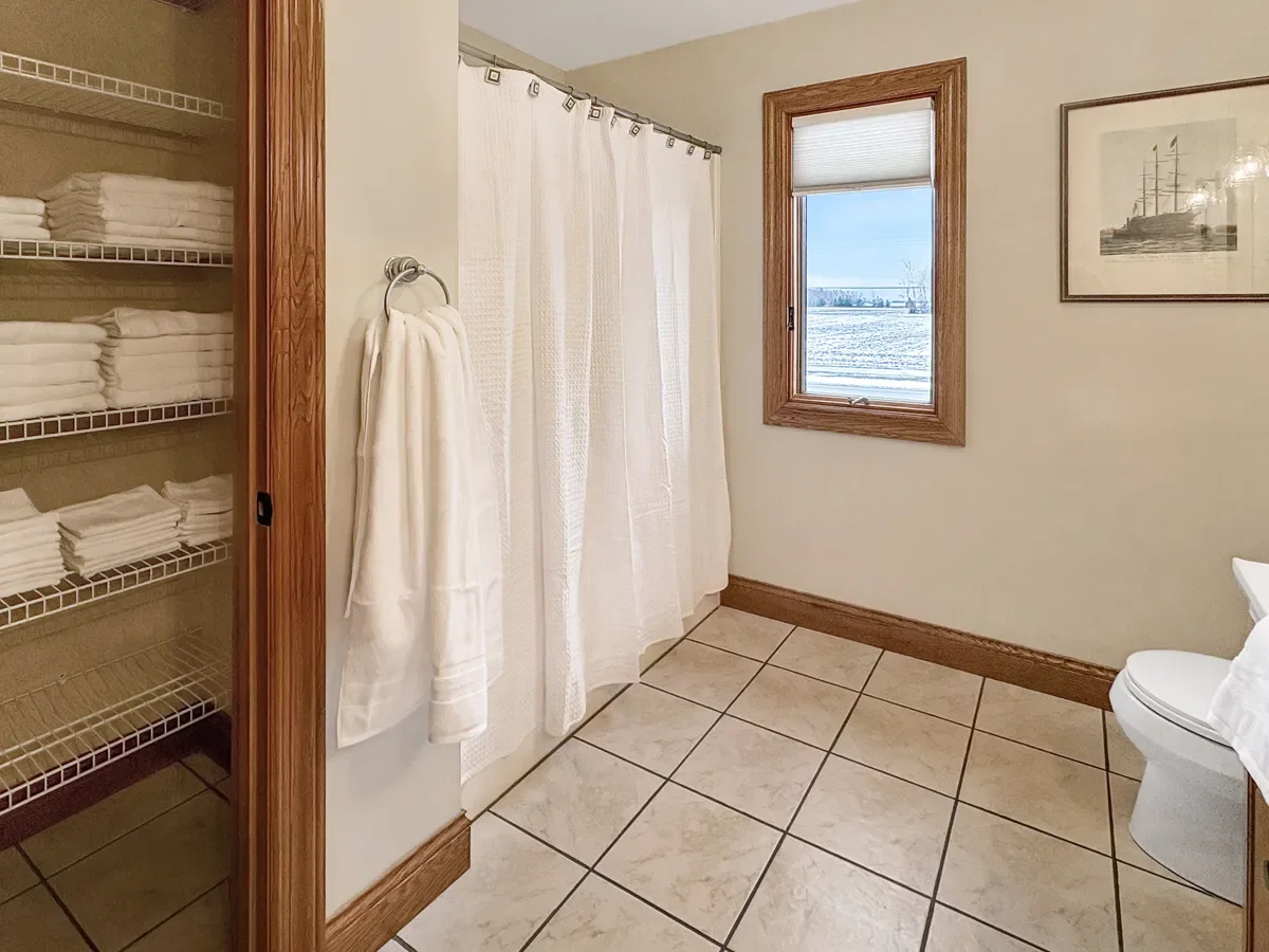 A bathroom with beige tiled floor, a window with a view of snow-covered ground, a white shower curtain, a towel hanging on the wall, a framed picture of a ship, and a bathroom shelf filled with folded towels.