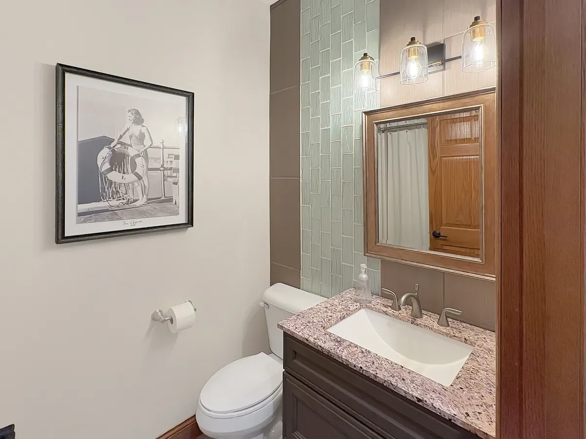 Bathroom with a framed black and white artwork of a woman on the wall, a toilet, a granite countertop with a sink, a soap dispenser, a wooden mirror cabinet, and modern light fixtures.