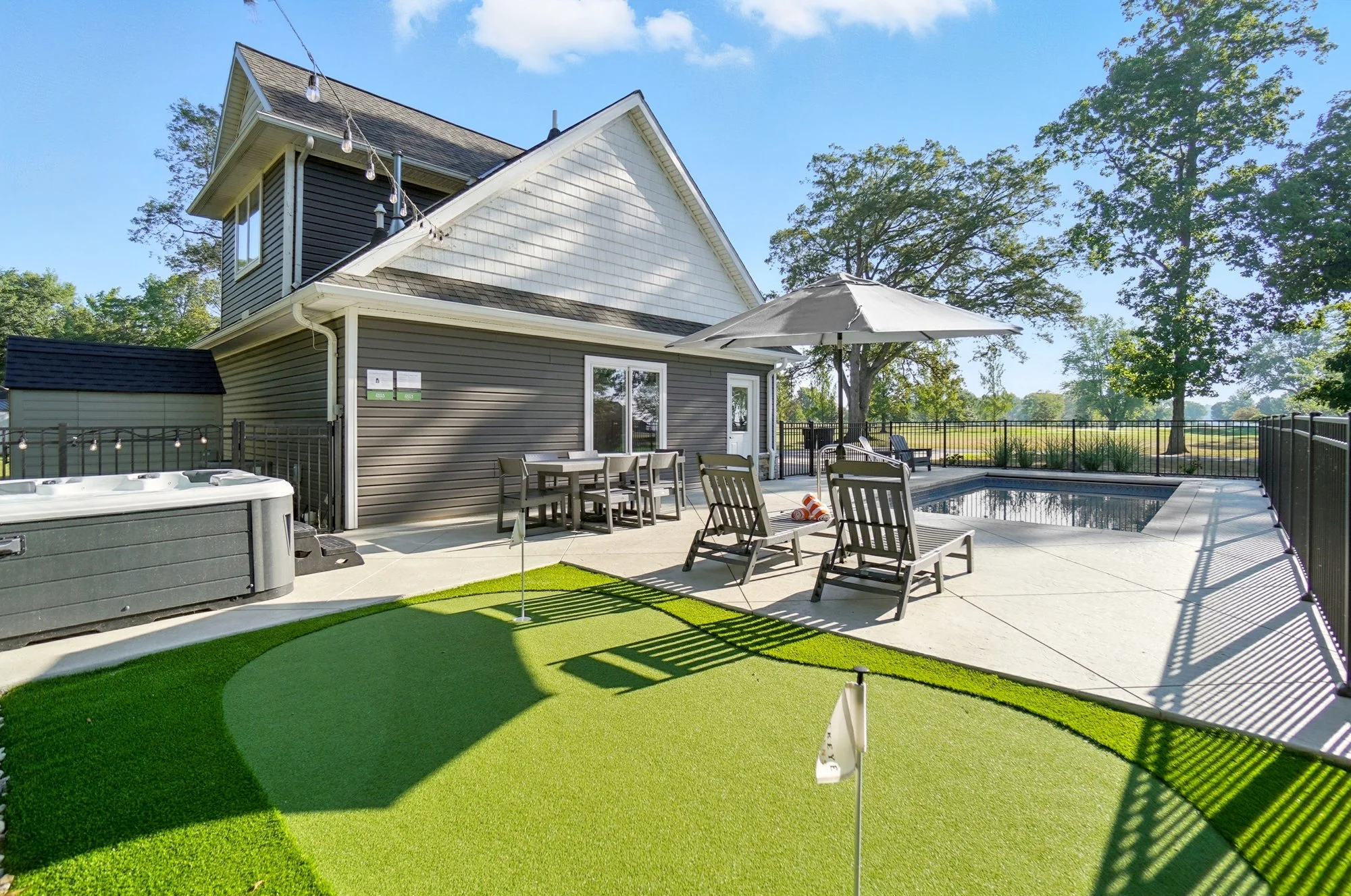 Backyard with hot tub, outdoor dining table, lounge chairs with umbrella, swimming pool, and artificial turf putting green, surrounded by a black fence, with trees and a field in the background under a clear blue sky.