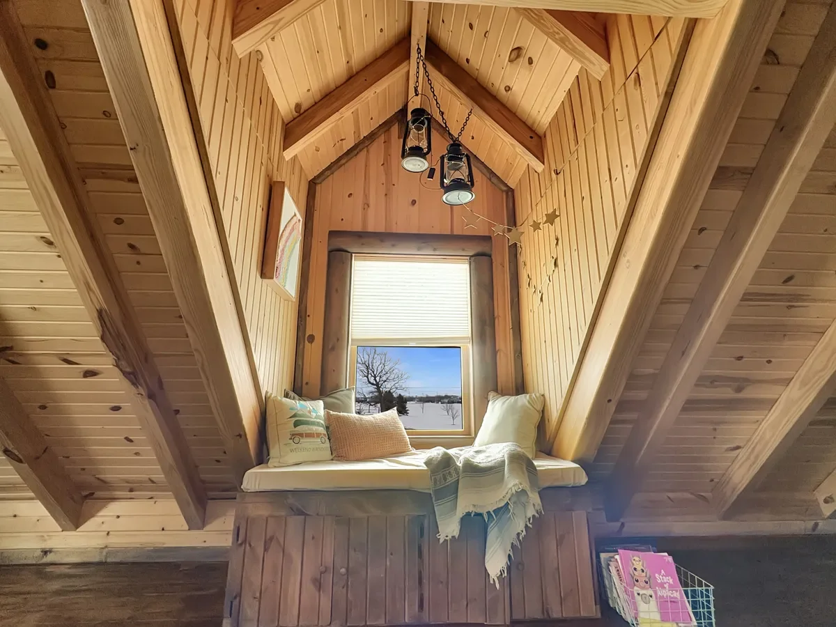 A cozy window seat with pillows and a blanket in a wood-paneled attic space, featuring a window with a snowy landscape outside, a ceiling with lantern-style lights, and star-shaped decorations.