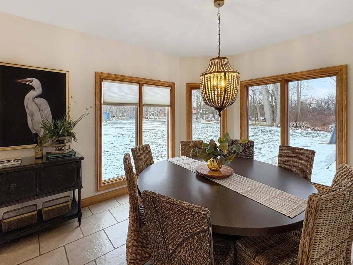 A dining room with a dark oval table, six wicker chairs, a hanging light fixture, a black sideboard with decorative plant, and five large windows showing a snowy outdoor landscape.