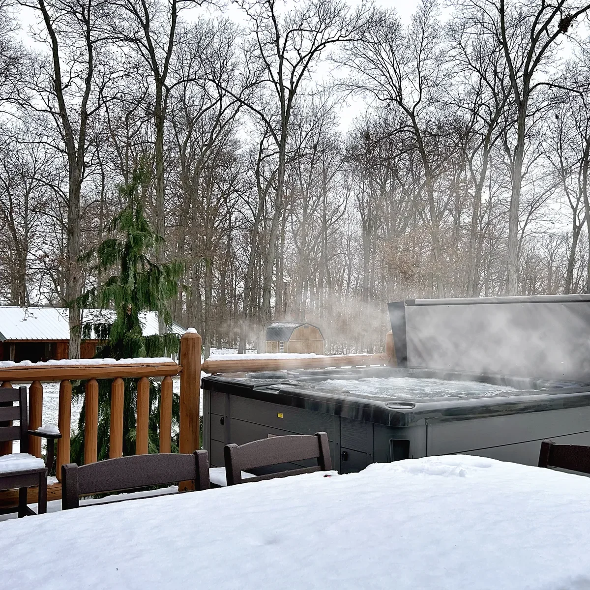 A snow-covered outdoor hot tub on a wooden deck, with steam rising from the hot water. Snow covers the furniture and surrounds a forest of leafless trees and a small shed in the background.
