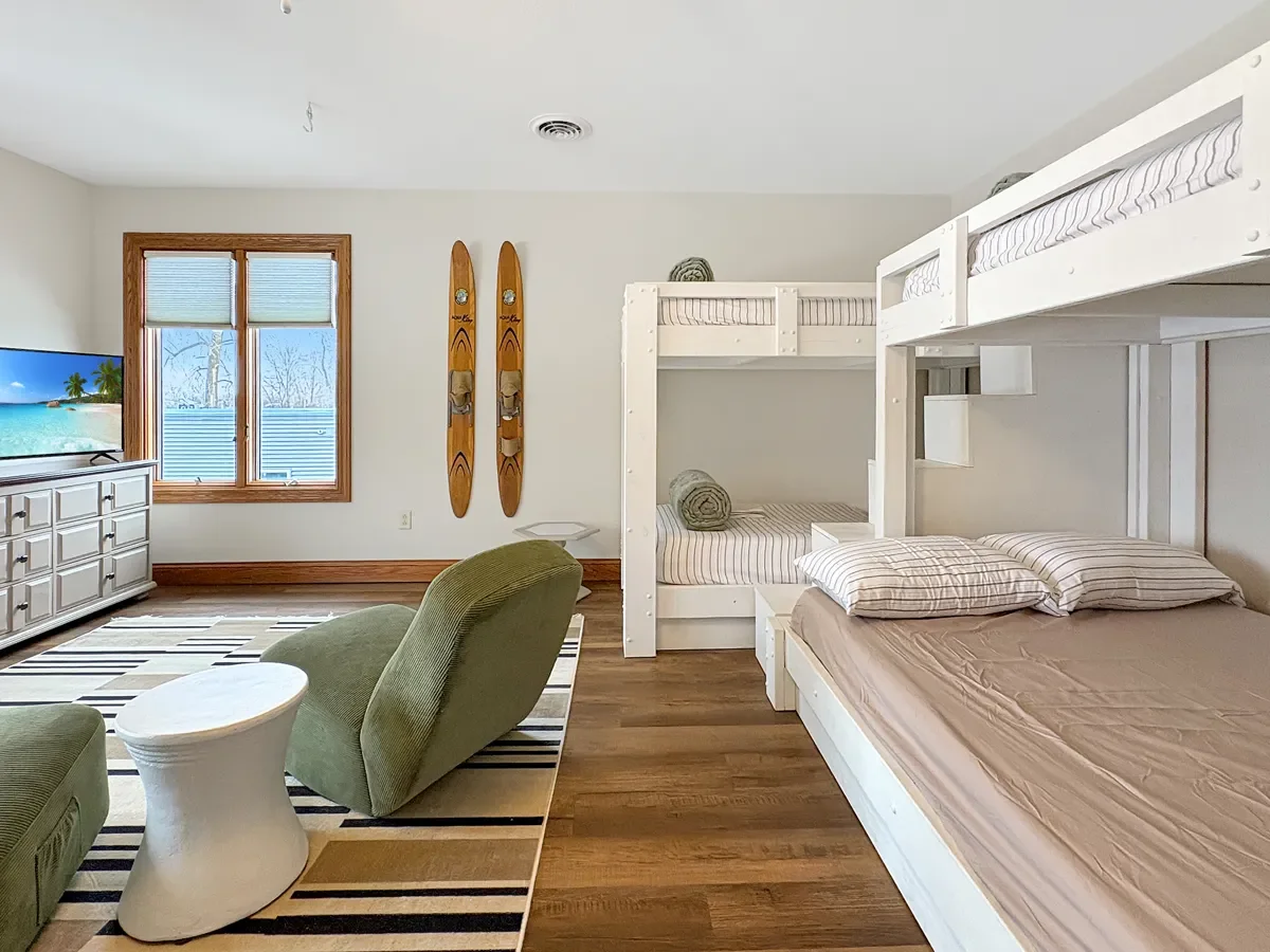 Room with two bunk beds, a green armchair, a white side table, a striped rug, a window with wooden frame, a white dresser, and a flat-screen TV showing a beach scene.