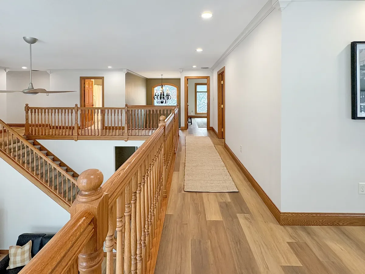 Interior view of a home with hardwood flooring, white walls, a staircase with wooden banister, and a hallway leading to various rooms. A chandelier hangs near a large window at the end of the hallway.