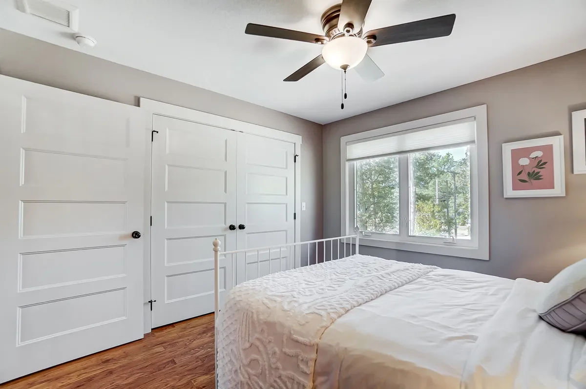 A bedroom with a white metal bed frame, a closed white closet door, a large window with a view of trees outside, and framed art on the wall. There is a ceiling fan with a light fixture and wooden flooring.