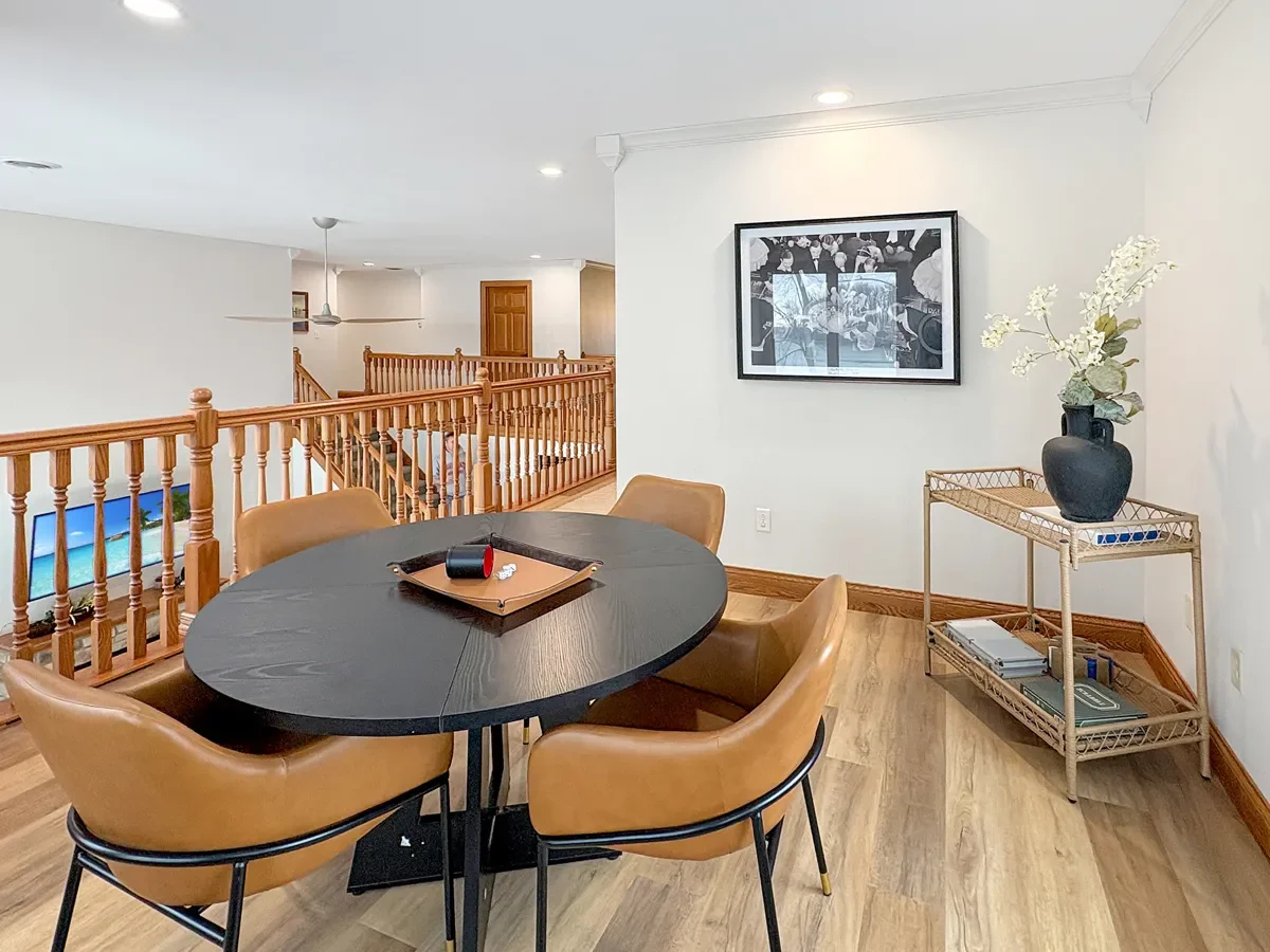 Dining area with a black round table and four brown chairs, a wall with framed black and white artwork, a beige corner table with a black vase of white flowers, and wooden railing overlooking a staircase.