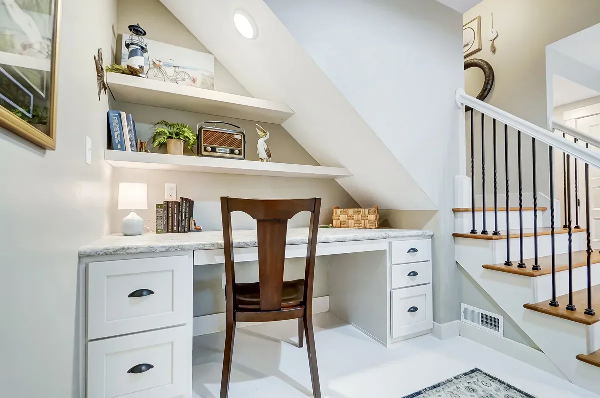 Home office under staircase with built-in desk, bookshelf, decorative items, and a chair.